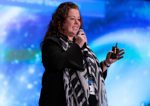 we-are-5 Speaker presenting on stage with microphone against blue background, wearing black and white patterned scarf.