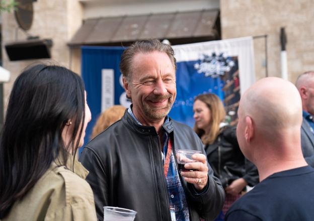 we-are-4 People socializing at an outdoor gathering, with a person in a black leather jacket holding a drink and smiling.