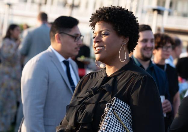 we-are-3 Person with curly hair wearing black outfit and hoop earrings at outdoor event with blurred crowd in background.