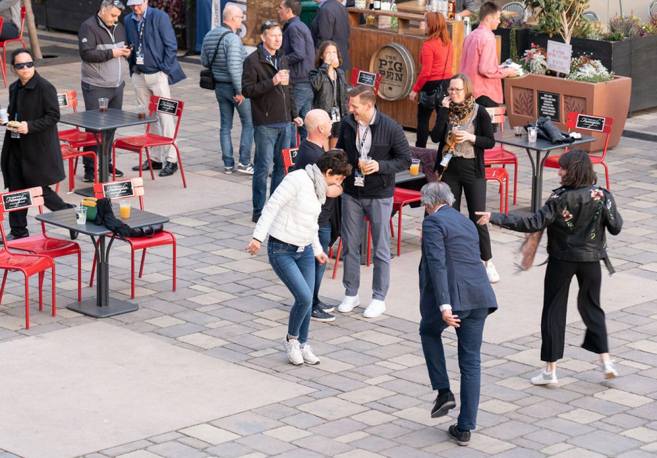 we-are-6-1 People socializing at outdoor café with red chairs, enjoying drinks near a wooden barrel marked "The Pig Pen"