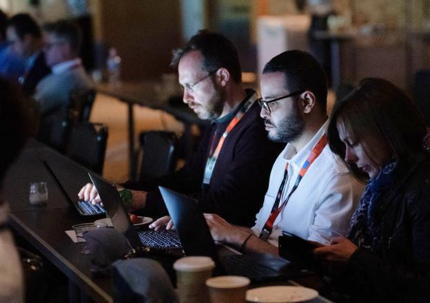 we-are-2 Attendees with conference badges working on laptops in a dimly lit auditorium with coffee cups on the table.