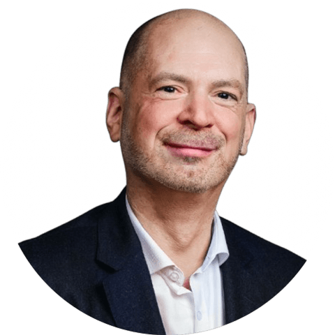 Bald man with stubble smiling in professional headshot, wearing dark blazer and white shirt against white background.