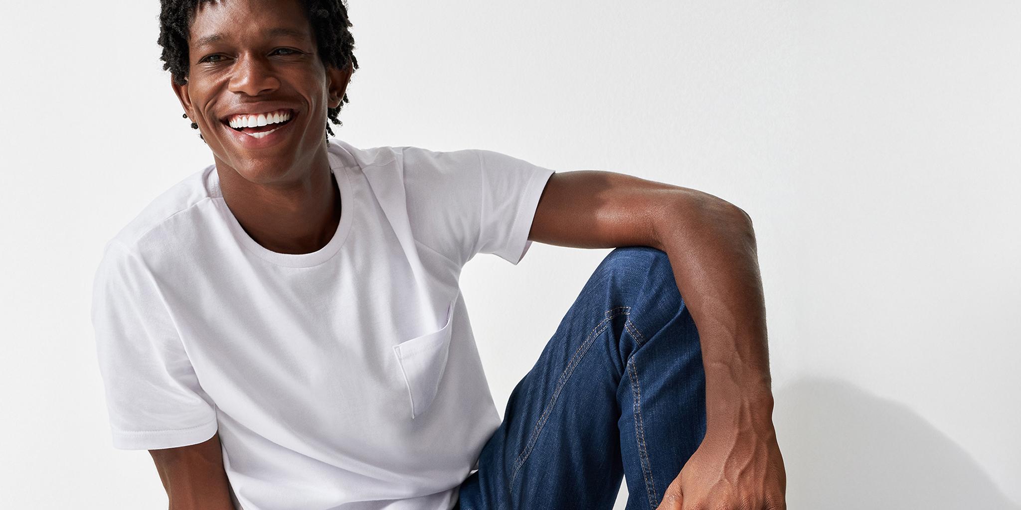 Malwee Group Enhances Expertise by Replacing Proprietary Software with Centric PLM Featured A young man with short curly hair smiles widely while sitting against a white wall. He is wearing a white t-shirt and blue jeans, appearing relaxed and cheerful—like someone enjoying the ease of Centric PLM’s proprietary software.