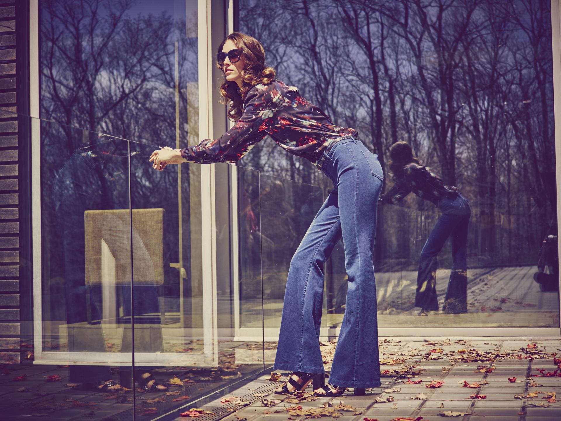 One Jeanswear Group chooses Centric 8 PLM Product Lifecycle Management (PLM) to be used across all brands Featured A woman in flared jeans and a floral blouse from One Jeanswear Group is leaning on a glass railing. She is wearing sunglasses and standing next to a large glass door reflecting her image. The outdoor setting, filled with dry leaves and bare trees, suggests late autumn or early winter.