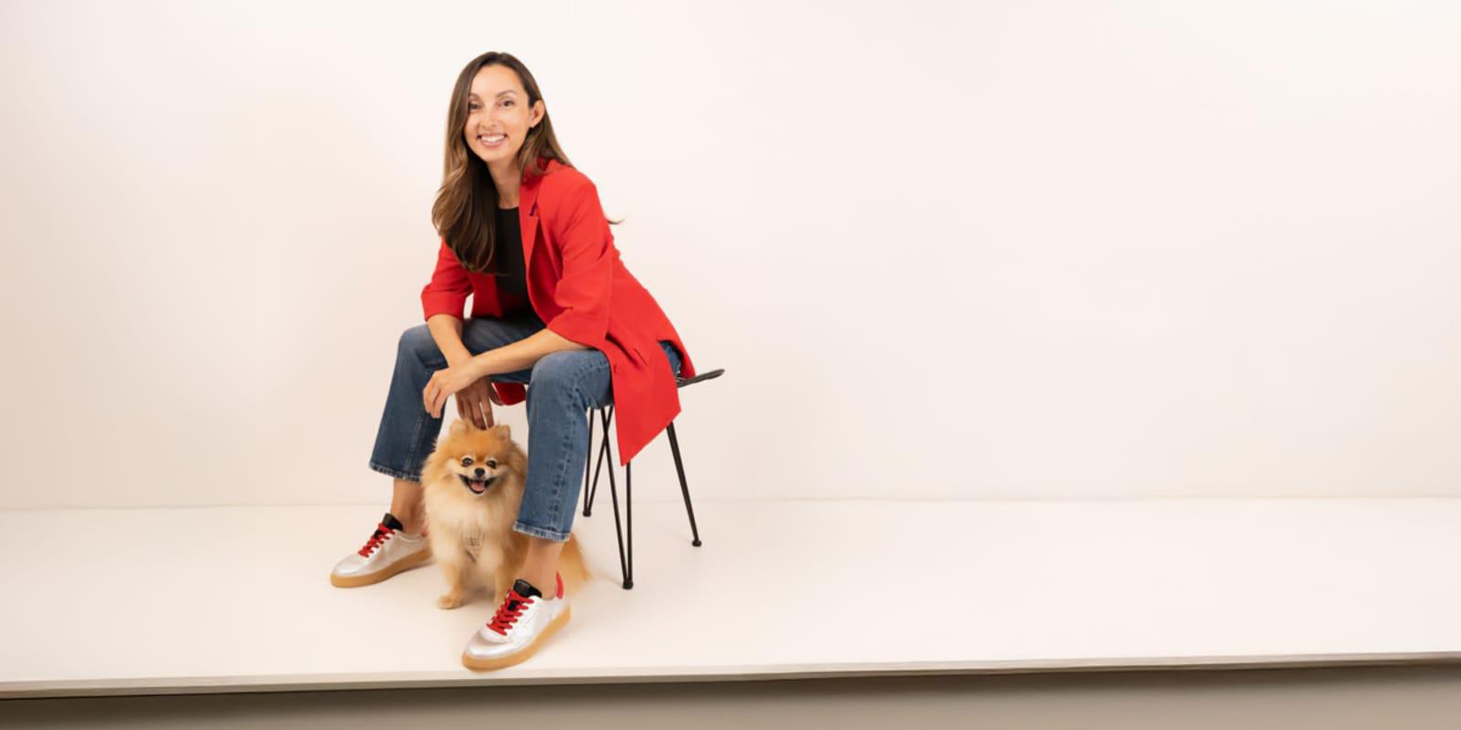 GLDN by Görtz Steps Up Product Development with Centric PLM Featured Một người phụ nữ mỉm cười trong chiếc áo khoác đỏ và quần jeans ngồi trên ghế, một tay đặt lên chú chó nhỏ lông xù dưới chân. Nền trắng đơn giản làm nổi bật phong cách tự tin của cô, gợi nhớ đến tinh thần đổi mới đằng sau quá trình phát triển sản phẩm GLDN by Görtz.