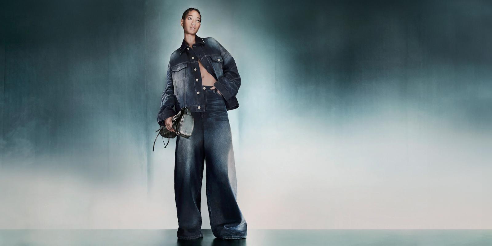 Acne Studios Supports Growth Ambitions with Centric Planning Featured A person poses confidently in an oversized denim jacket from Acne Studios and wide-leg jeans against a misty gradient background. The individual holds a stylish handbag, with one hand casually placed in a jacket pocket. The overall mood is chic and modern, embodying growth ambitions in fashion.