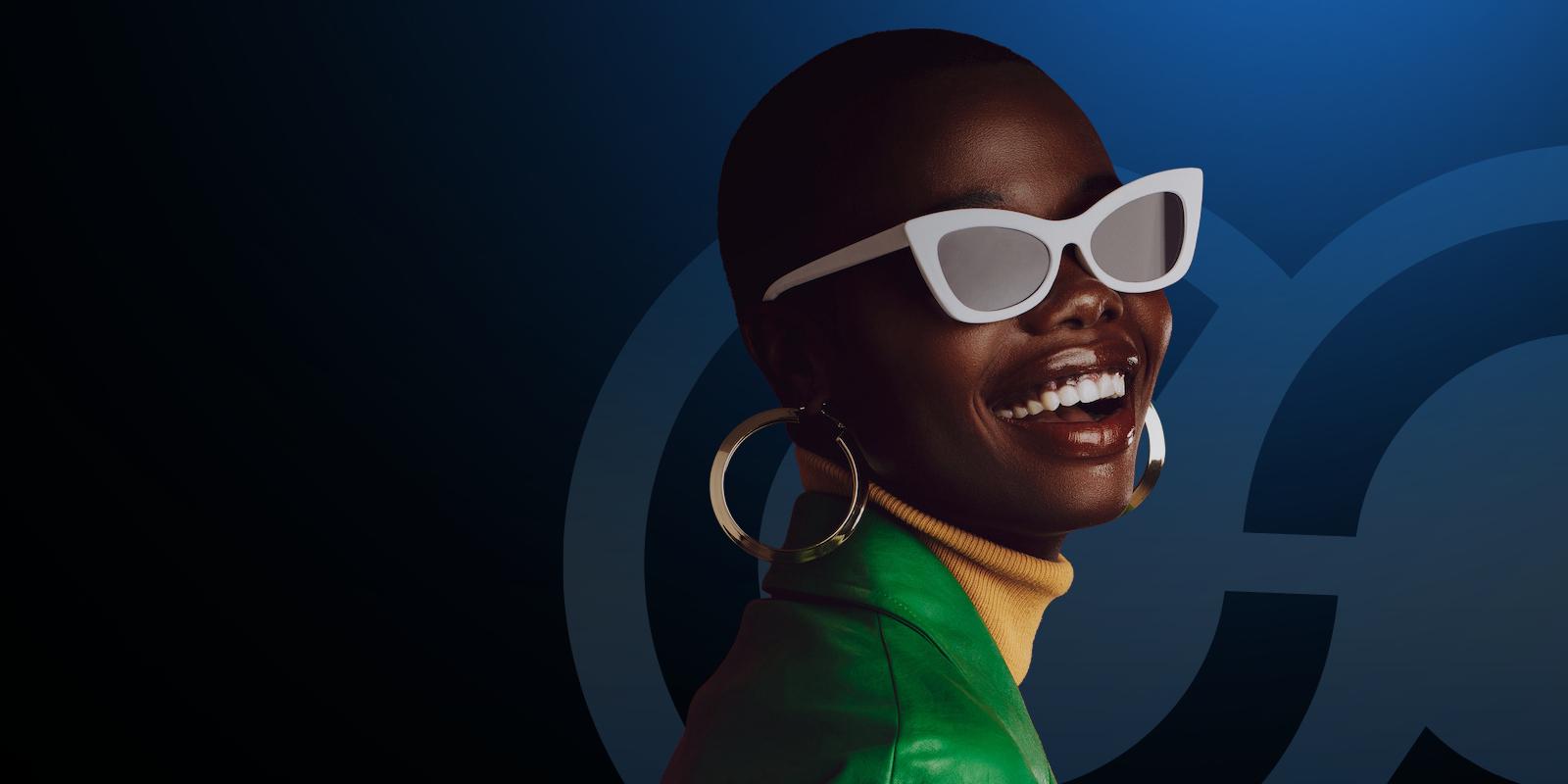 FEBRATEX SUMMIT 2025 Feature Uma pessoa sorridente com pele escura usa óculos de sol brancos, brincos de argola grandes, uma camisola de gola alta amarela e um casaco verde sobre um fundo azul escuro com padrões circulares.