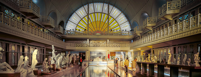 The interior of a grand museum, ideal for a Séminaire PLM, boasts an arched, stained-glass window featuring a sunburst design. The space is adorned with classical sculptures on both sides of the central aisle, illuminated by warm ambient lighting. High ceilings and two-level balconies enhance its regal atmosphere.