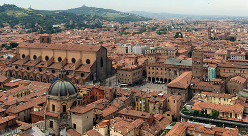 An aerial view of Bologna, Italy, centric for small business, showcases its terracotta rooftops, medieval buildings, and historic towers. Dominating the scene is the large Basilica of San Petronio and Piazza Maggiore, nestled among densely packed, picturesque architecture.