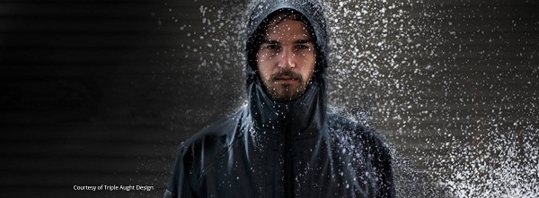 A person stands facing the camera, wearing a dark, hooded jacket. Water droplets spray around him, highlighting the jacket's water-resistant properties. The background is dimly lit, creating a dramatic effect with the illuminated water droplets—perfect for small businesses aiming to stay competitive in outerwear.
