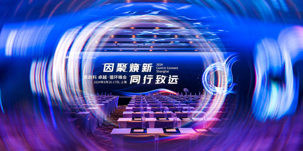 A conference room set up for the "2024 Centric Connect Shanghai" event. The room features rows of tables and chairs, each table with a notepad and pen. The backdrop displays the event's name in Chinese and English with circular graphics overlaying the image, highlighting Centric PLM's role in addressing supply chain disruptions.