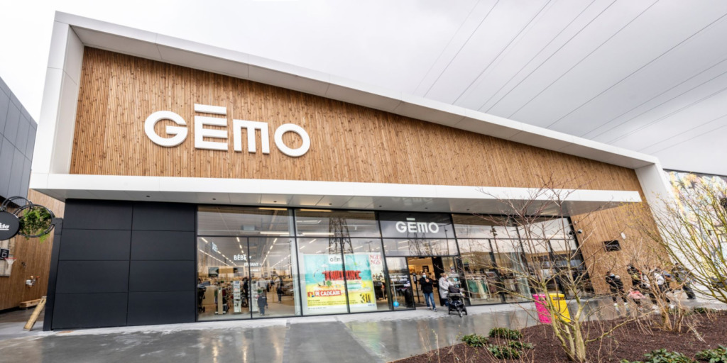 A modern retail store with a wooden facade and large glass windows under a sign that reads "GÉMO" showcases its commitment to global transformation. The store entry is bustling with people walking in and out. The ground is wet, indicating recent or ongoing rain, with trees and shrubs visible in the foreground.