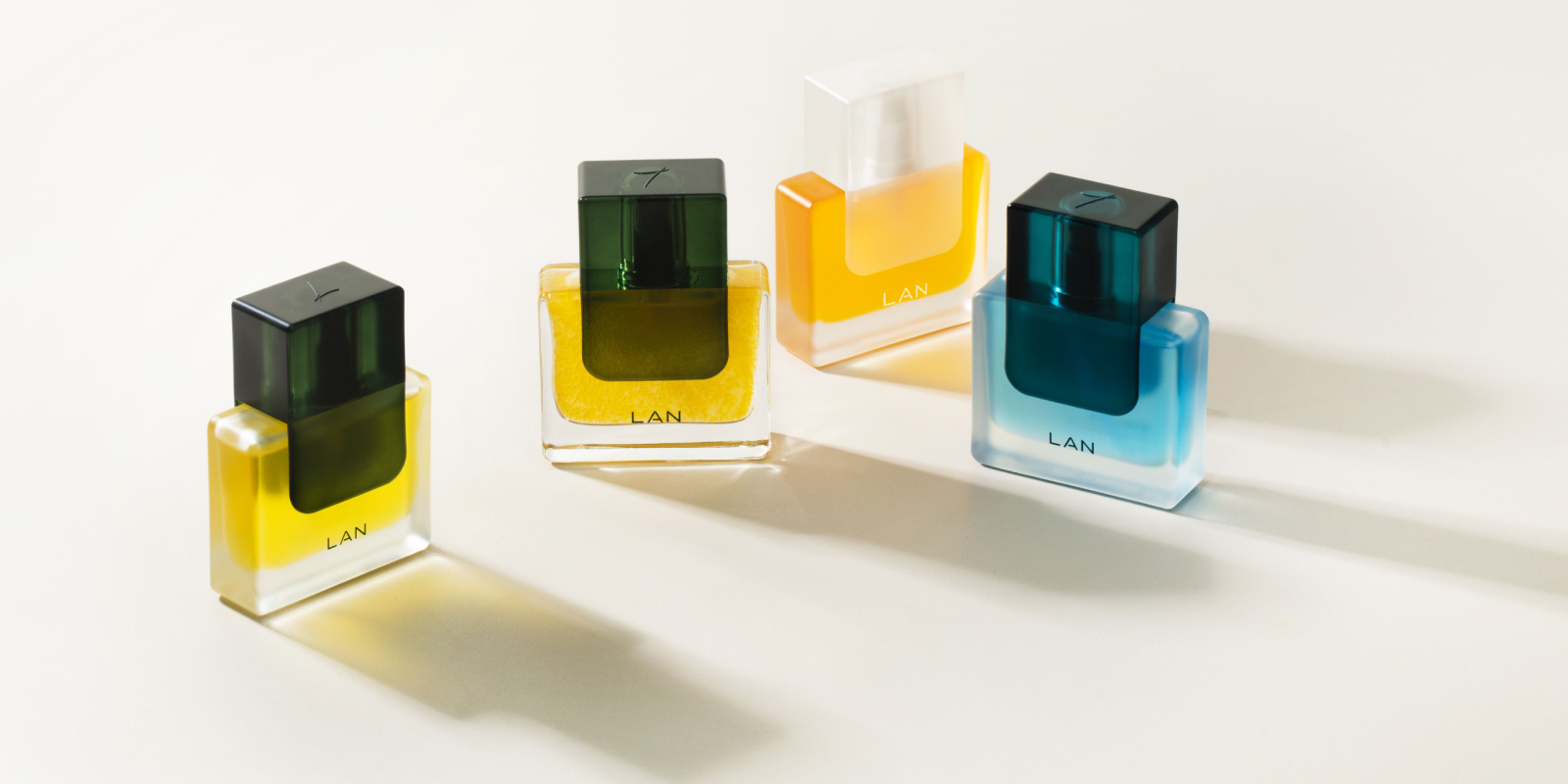 Sobre una superficie clara se muestran cuatro frascos de perfume con la etiqueta "LAN". Cada frasco presenta una combinación de colores única: verde-amarillo, amarillo-transparente, verde-azul y azul-azul claro. Los frascos proyectan sombras suaves sobre la superficie.
