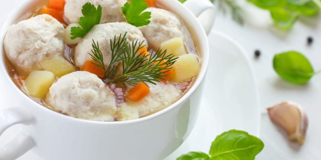 A white bowl filled with soup containing meatballs, diced carrots, and potatoes. Garnished with fresh parsley and dill. In the background, there's garlic, peppercorns, and green leaves. The setting is bright and inviting.