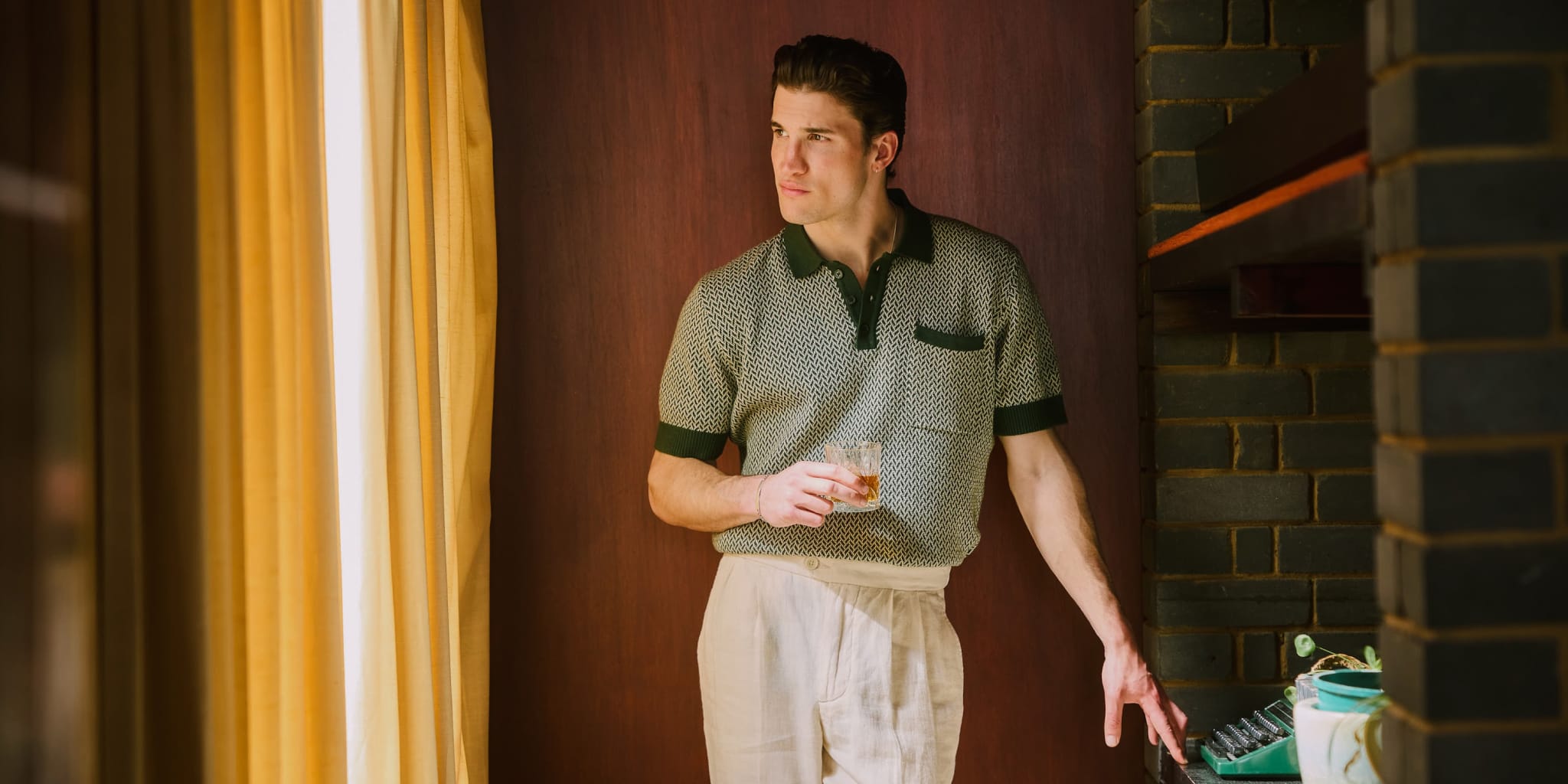 Un uomo con una polo a fantasia e pantaloni chiari si trova al chiuso, accanto a una finestra con tende gialle, con in mano un drink e lo sguardo rivolto all'esterno, mentre l'altra mano è appoggiata a un muro di mattoni scuri.