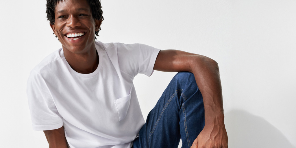Una persona sorridente che indossa una maglietta bianca e jeans blu siede con disinvoltura su uno sfondo bianco.